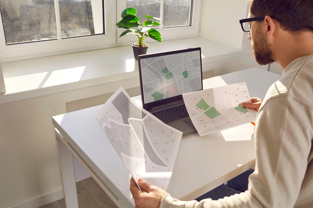 serious engineer with cadastral maps and projects is sitting at desk with laptop in office. cadastral engineer carries out accounting of land plots. ownership of real estate object.